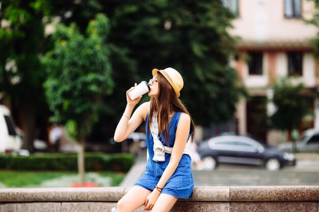 young woman wearing denim dress with straw hat drinking coffee